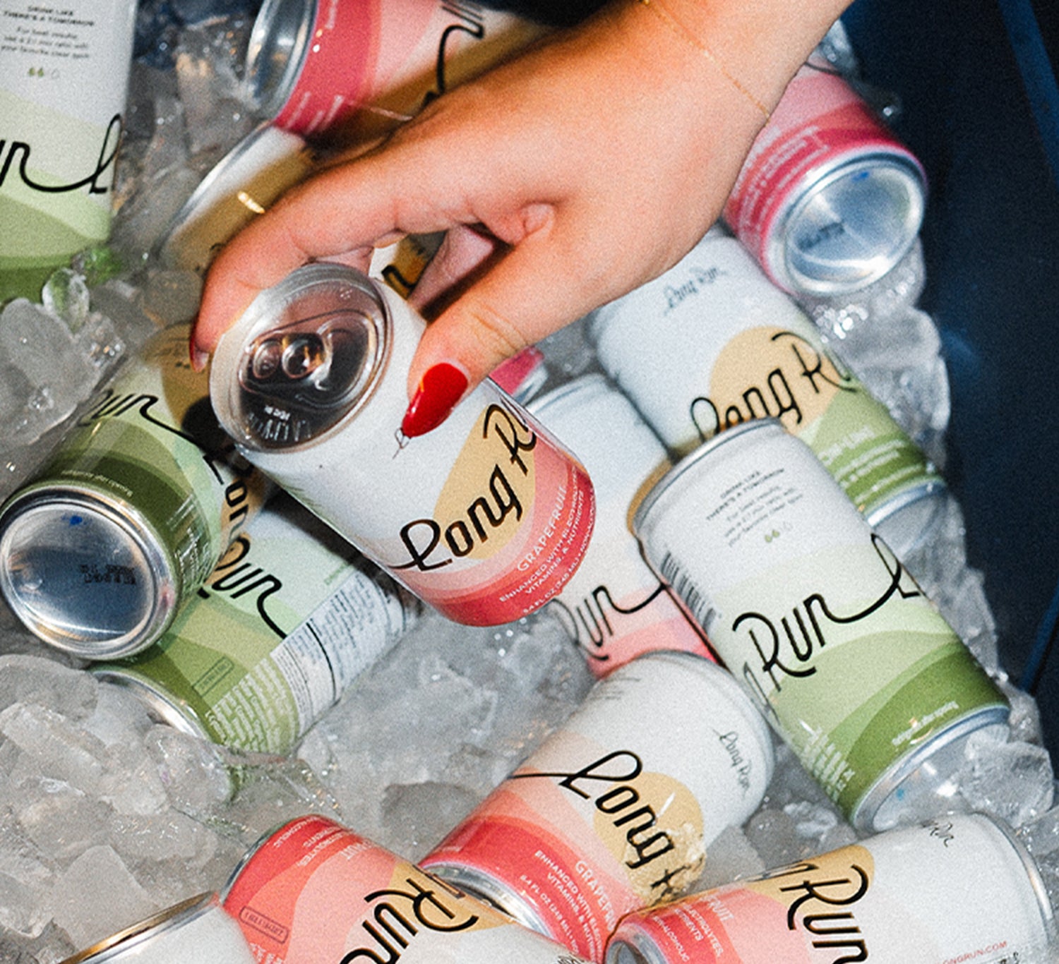 A hand with red nail polish reaches for a can of Long Pine Rum from an assortment of other Long Pine Rum cans, some with pink and green labels, resting in a bed of ice.