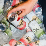 A hand with red nail polish reaches for a can of Long Pine Rum from an assortment of other Long Pine Rum cans, some with pink and green labels, resting in a bed of ice.