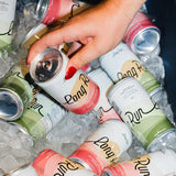 A hand with red nail polish reaches for a can of Long Pine Rum from an assortment of other Long Pine Rum cans, some with pink and green labels, resting in a bed of ice.