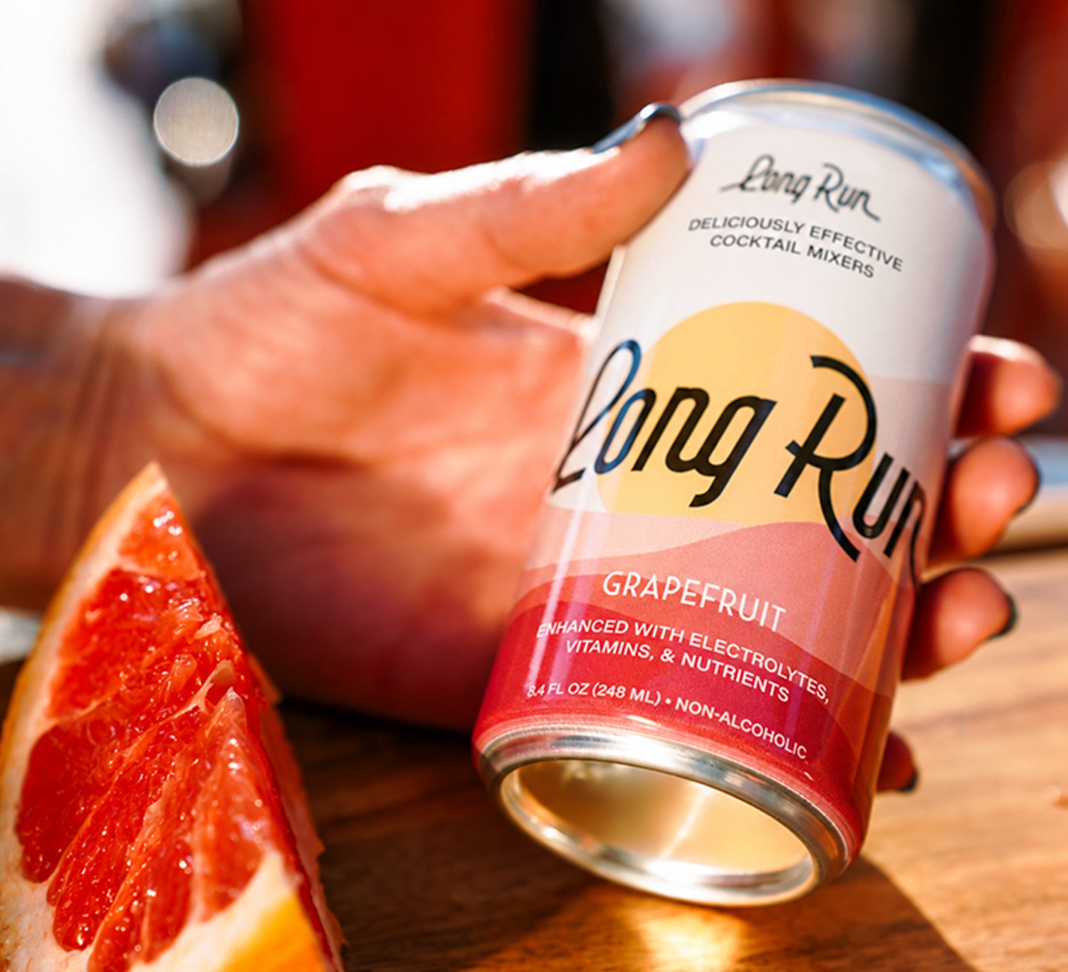 A hand holds a Long Run Grapefruit cocktail mixer  in a warm, sunny setting next to a grapefruit slice on a wooden surface.
