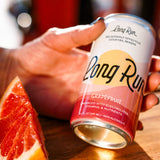 A hand holds a Long Run Grapefruit cocktail mixer  in a warm, sunny setting next to a grapefruit slice on a wooden surface.